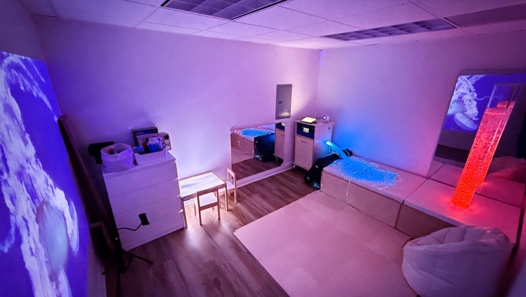 Sensory Bridge sensory room with colored lighting, a bubble tube, fiber-optic strands, mirrors, and a padded seating area.