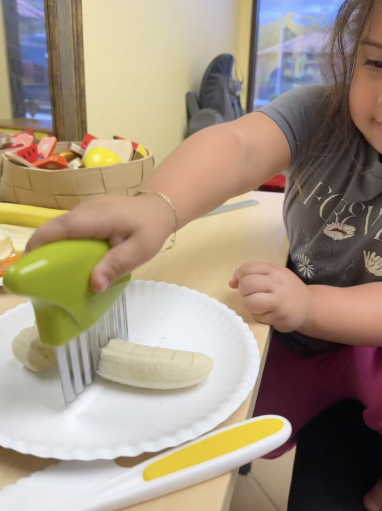 Child cutting a banana with a green handled food cutter at a table.