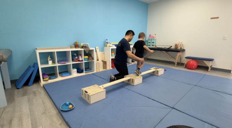 Adult supporting a child walking along a wooden balance beam.